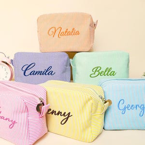 Puede incluir: Varias bolsas de cosméticos a rayas en tonos rosa, azul, amarillo y naranja, cada una bordada con un nombre. Las bolsas tienen cierre de cremallera. Un despertador rosa es visible en el fondo. Ideales para guardar maquillaje y artículos de aseo.
