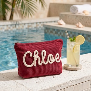 May include: A red corduroy cosmetic bag with the name "Chloe" embroidered in white rope-like letters. The bag has a gold zipper and is next to a glass of lemonade with a straw and lemon slices.
