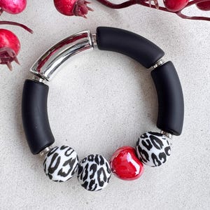 Könnte beinhalten: Ein Armband mit schwarzen und weißen Perlen mit Leopardenmuster und einer glänzenden roten Perle. Das Armband hat eine silberfarbene, gebogene Stange und schwarze zylindrische Perlen. Ein modisches Accessoire.