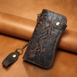 Puede incluir: Cartera de cuero marrón oscuro con un diseño de dragón en relieve. La cartera tiene un cierre de botón a presión y un llavero de cuero a juego. El dragón está detallado, agregando una textura única y atractivo visual.