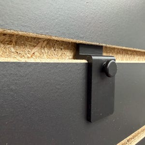 May include: Close-up of a black metal bracket with a round head screw, attached to a black wooden panel. The panel is made of particle board and has a visible wood grain texture.