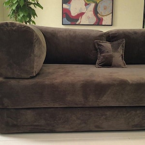 May include: A dark brown corduroy sofa featuring a low-profile design with a thick, cylindrical bolster armrest on the left side and a square, patchwork-style accent pillow resting on the seat cushion, all upholstered in a textured fabric.
