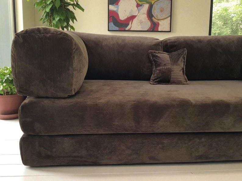 May include: A dark brown corduroy sofa featuring a low-profile design with a thick, cylindrical bolster armrest on the left side and a square, patchwork-style accent pillow resting on the seat cushion, all upholstered in a textured fabric.