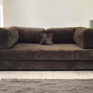 May include: A dark brown corduroy sofa featuring a low-profile design with thick, rounded armrests and a single square accent pillow placed in the center of the back cushions. The upholstery has a distinct ribbed texture throughout the entire frame.