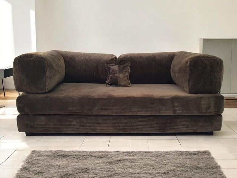 May include: A dark brown corduroy sofa featuring a low-profile design with thick, rounded armrests and a single square accent pillow placed in the center of the back cushions. The upholstery has a distinct ribbed texture throughout the entire frame.