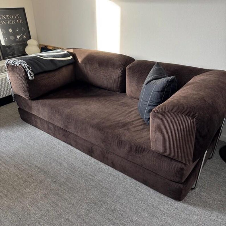 May include: A dark brown corduroy sofa featuring a low-profile design with thick, rectangular armrests and a deep seat. The sofa is accented with a black and white patterned throw blanket on the left side and a dark gray striped throw pillow.