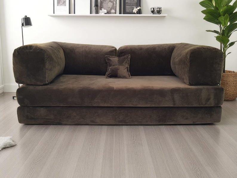 May include: A dark brown corduroy sofa featuring a low-profile design with thick, rectangular armrests and a matching square throw pillow centered on the back cushions. The upholstery has a distinct ribbed texture throughout the entire frame.