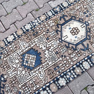 May include: A rectangular, patterned rug with a beige, blue, and white color scheme. The rug features geometric designs and a border with a repeating pattern. The rug is placed on a gray brick surface.