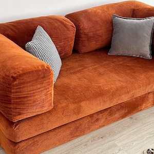 May include: A rust-colored corduroy sofa featuring a low-profile design with thick, rounded armrests and a metal frame base. The sofa includes one small black and white checkered throw pillow and one solid gray square throw pillow for accent.