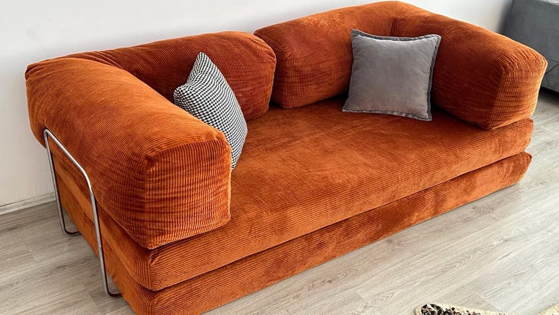 May include: A rust-colored corduroy sofa featuring a low-profile design with thick, rounded armrests and a metal frame base. The sofa includes one small black and white checkered throw pillow and one solid gray square throw pillow for accent.