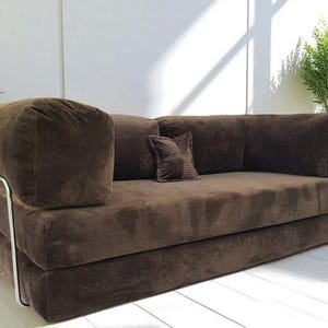 May include: A brown corduroy sofa featuring a low-profile design with thick, rectangular cushions and a single small square accent pillow. The sofa is supported by a sleek, silver-toned metal frame that wraps around the exterior sides.