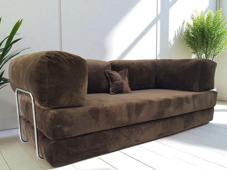 May include: A brown corduroy sofa featuring a low-profile design with thick, rectangular cushions and a single small square accent pillow. The sofa is supported by a sleek, silver-toned metal frame that wraps around the exterior sides.