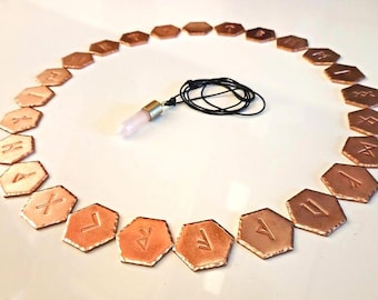 Puede incluir: Un conjunto de piedras rúnicas hexagonales de color cobre dispuestas en círculo, cada una grabada con un símbolo diferente. Un colgante de cristal de cuarzo rosa cuelga de un cordón negro en el centro. Las runas se utilizan probablemente para la adivinación.