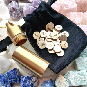 May include: A set of small hexagonal brass tokens engraved with runic symbols, accompanied by a matching cylindrical brass container with a screw-top lid and a black velvet drawstring pouch for storage and transport.
