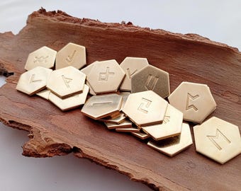Puede incluir: Una colección de piedras rúnicas hexagonales de color dorado con símbolos grabados. Las piedras están dispuestas sobre un trozo de corteza marrón texturizada. Las runas son de varios estilos, y la estética general es rústica y mística.