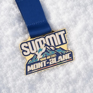 Peut inclure: Une médaille en métal doré avec un ruban bleu portant l'inscription SUMMIT MONT-BLANC et 4806 METERS. Le design comprend un graphique de montagne stylisé en émail bleu, blanc et turquoise, avec une bordure bleu foncé et un ruban en tissu bleu.
