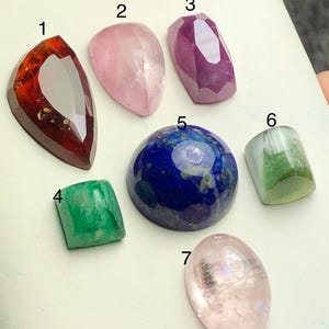 Puede incluir: Una colección de siete piedras preciosas de varias formas y colores. Las piedras preciosas incluyen piedras de color rojo oscuro, rosa, morado, azul, verde y transparente. Las piedras se exhiben sobre una superficie clara.