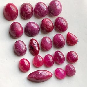 May include: A collection of polished, oval-shaped gemstones in varying shades of red and pink. The gemstones are arranged on a white surface, with a teardrop-shaped stone and a long, boat-shaped stone among them.