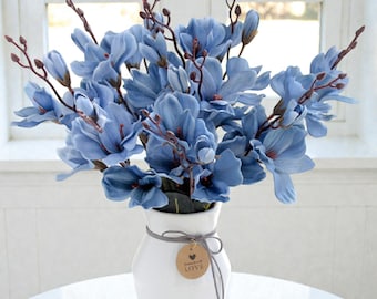Könnte beinhalten: Ein Strauß blauer Magnolienblüten mit braunen Stielen in einer weißen Keramikvase. Um den Hals der Vase ist eine graue Schnur gebunden, an der ein rundes braunes Papieretikett mit einem Herzsymbol und dem aufgedruckten Wort LOVE hängt.