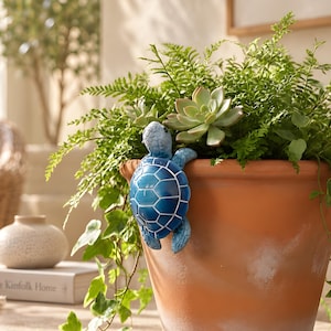 May include: A blue ceramic turtle figurine with a white shell pattern hangs over the rim of a large terracotta plant pot. The pot contains a lush green fern and a succulent, with trailing ivy leaves cascading down the side of the orange clay vessel.