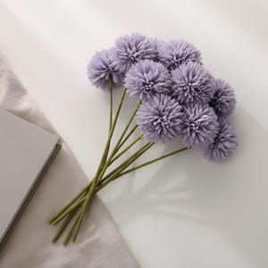 May include: A collection of artificial lavender pom-pom flowers with green stems. The flowers are bunched together, giving a full, textured look. The stems are long and thin, and the flowers are arranged on a white surface with a light grey cloth.