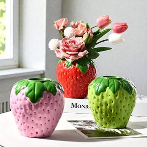 Puede incluir: Tres jarrones de cerámica con forma de fresa en rojo, rosa y verde. El jarrón rojo contiene un ramo de rosas y tulipanes rosas. Los jarrones tienen tapas verdes con forma de hoja y pequeños detalles de puntos, ideales para la decoración del hogar.