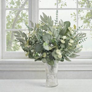 May include: A vibrant arrangement of artificial eucalyptus and other greenery in a clear, textured glass vase. The display features various shades of green leaves and small white flowers, creating a natural and elegant look. The vase is on a white surface in front of a window.