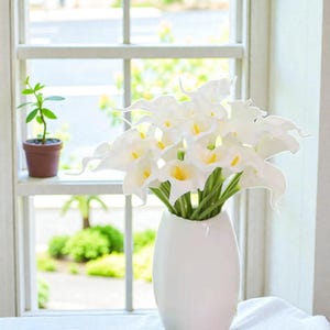 May include: A white ceramic vase holds a bouquet of white calla lilies with yellow centers and green stems. The vase sits on a white surface near a window with a small potted plant.