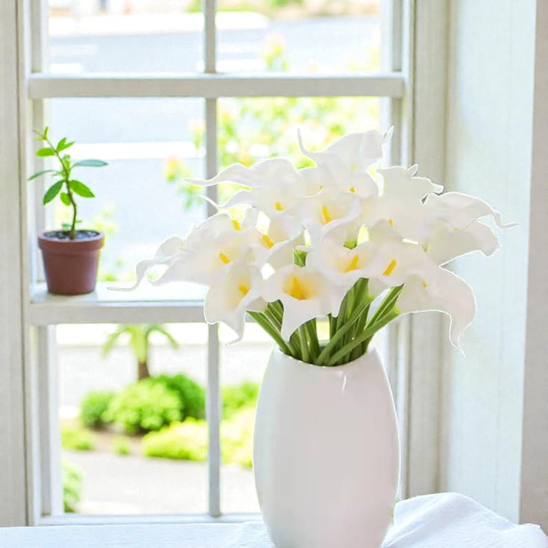 May include: A white ceramic vase holds a bouquet of white calla lilies with yellow centers and green stems. The vase sits on a white surface near a window with a small potted plant.