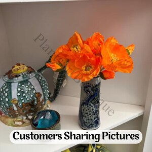 K&ouml;nnte beinhalten: Ein Strau&szlig; leuchtend orangefarbener Mohnblumen in einer hohen, schlanken, grau und blau gesprenkelten Keramikvase. Neben der Vase steht eine runde, mehrfarbige Keramikteekanne mit strukturierter Oberfl&auml;che und eine kleine Schale mit blauen Glassteinen.