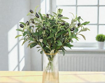 May include: A glass vase with a gold rim holds a bouquet of olive branches. The branches have green leaves and dark olives. The vase is clear with vertical ridges and sits on a light-colored wooden surface.
