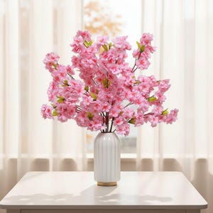 May include: A white ceramic vase with vertical ridges holds a large arrangement of pink cherry blossom branches. The flowers have multiple layers of petals and green leaves. The vase sits on a white table with a gold base.