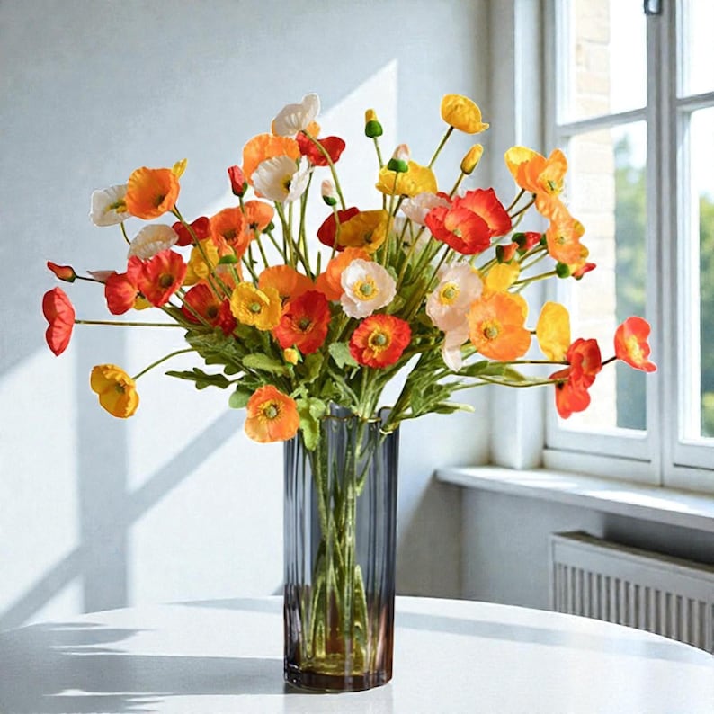 May include: A colourful bouquet of poppies in a tall, fluted glass vase. The flowers display shades of red, orange, yellow, and white, with green stems and leaves. The vase sits on a white surface near a window.