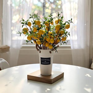 以下が含まれることがあります： ロープの持ち手が付いた白い金属製のバケツに、人工的なレモンの枝が詰められています。レモンは鮮やかな黄色で、葉は緑色です。バケツは木製の正方形の上に置かれ、前面には「FLOWERS & GARDEN」の文字があります。