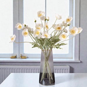 K&ouml;nnte beinhalten: Ein Strau&szlig; wei&szlig;er Mohnblumen mit gelben Mitten und gr&uuml;nen Stielen in einer hohen, durchscheinenden, rauchfarbenen Glasvase. Die Blumen haben zarte, d&uuml;nne Bl&uuml;tenbl&auml;tter und sind mit mehreren gr&uuml;nen Bl&auml;ttern an den Stielen arrangiert.