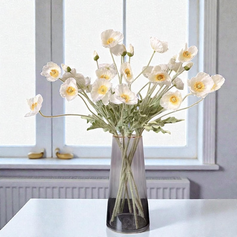 K&ouml;nnte beinhalten: Ein Strau&szlig; wei&szlig;er Mohnblumen mit gelben Mitten und gr&uuml;nen Stielen in einer hohen, durchscheinenden, rauchfarbenen Glasvase. Die Blumen haben zarte, d&uuml;nne Bl&uuml;tenbl&auml;tter und sind mit mehreren gr&uuml;nen Bl&auml;ttern an den Stielen arrangiert.