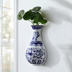 May include: A white and blue ceramic wall vase with a floral design, holding green leafy plants. The vase is mounted on a white wall. The vase has a curved shape and features intricate blue patterns.