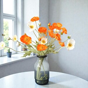 K&ouml;nnte beinhalten: Ein Strau&szlig; k&uuml;nstlicher Mohnblumen in leuchtendem Orange und klarem Wei&szlig; mit zarten Bl&uuml;tenbl&auml;ttern und gr&uuml;nen Stielen. Die Blumen sind in einer transparenten, zylindrischen Glasvase mit einem dicken Boden arrangiert.