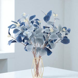 May include: A decorative arrangement of faux eucalyptus leaves in shades of blue and grey, with small white flower accents. The leaves are in a gold geometric vase, suitable for home decor.