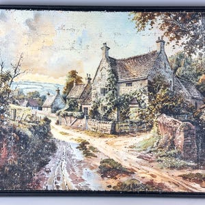 May include: Framed landscape painting depicting a quaint village scene. The artwork features a stone house with a thatched roof, surrounded by lush greenery and a dirt road. The color palette includes muted greens, browns, and blues, creating a serene atmosphere.