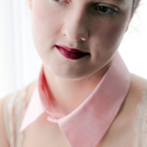 May include: A close-up of a woman's face with a pink collar. She has dark hair and is wearing a light pink collar with a pointed edge. She has a nose ring and dark red lipstick.