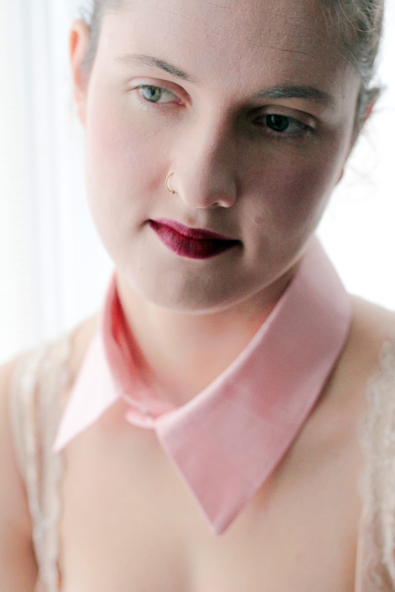 May include: A close-up of a woman's face with a pink collar. She has dark hair and is wearing a light pink collar with a pointed edge. She has a nose ring and dark red lipstick.