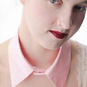 May include: A woman with a pink collared shirt and a nose ring. She is wearing dark red lipstick.
