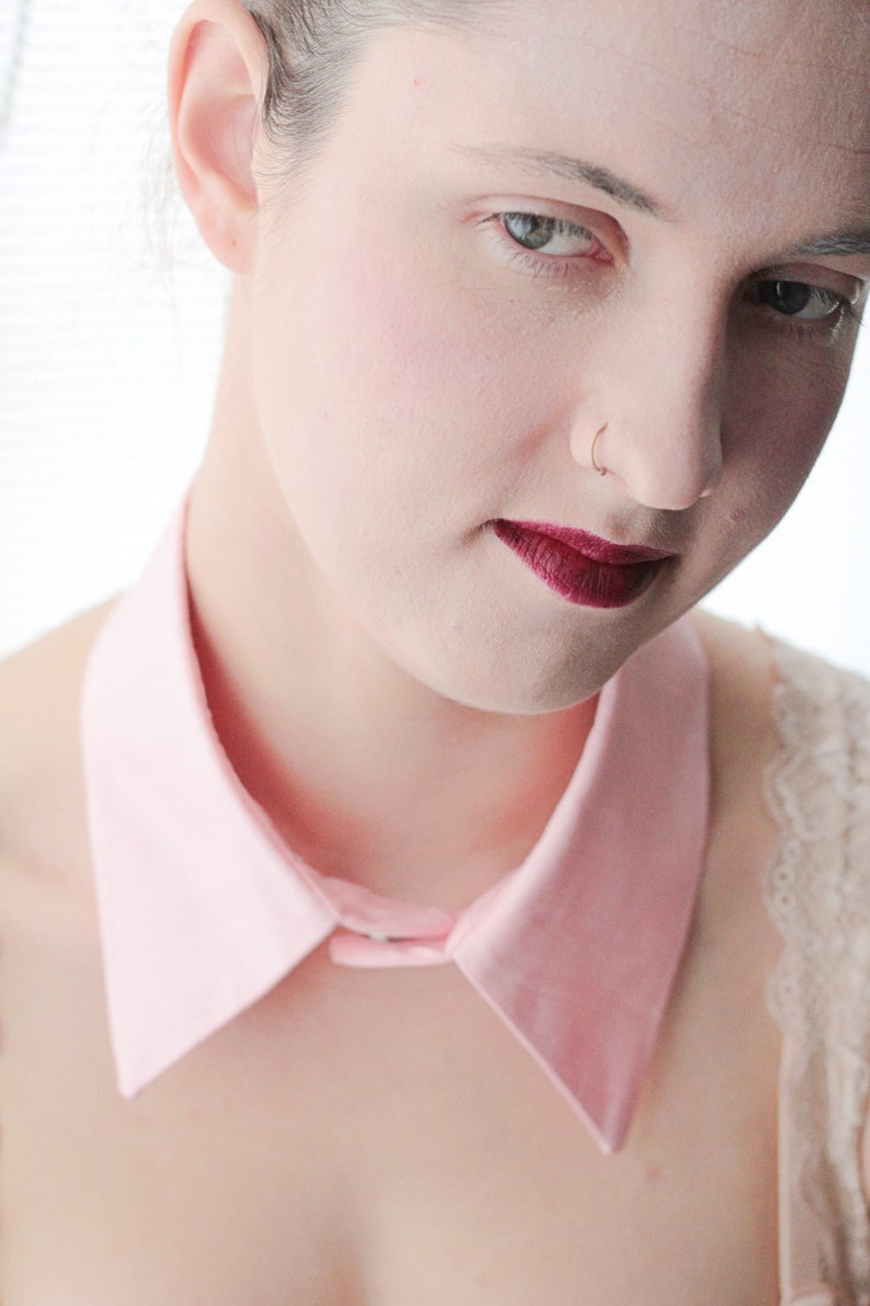 May include: A woman with a pink collared shirt and a nose ring. She is wearing dark red lipstick.