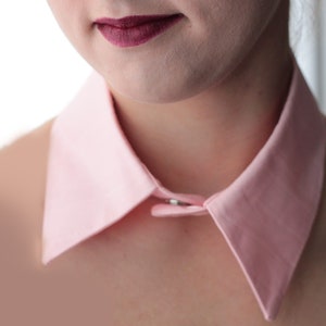 May include: A pink collared shirt with a button closure. The collar is large and stands up around the neck.