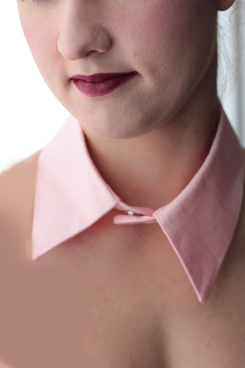 May include: A pink collared shirt with a button closure. The collar is large and stands up around the neck.