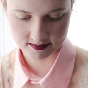 May include: A woman with a pink collar and a dark red lipstick is looking down. She is wearing a white lace top.