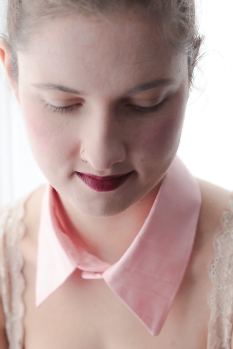 May include: A woman with a pink collar and a dark red lipstick is looking down. She is wearing a white lace top.