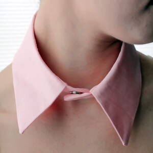 May include: A pink fabric collar with a silver button closure. The collar is folded down and is worn around the neck.