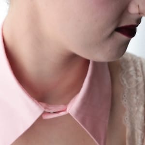 May include: A close-up of a person wearing a pink collared shirt with a button closure. The collar is large and has a pointed shape. The person is wearing a white lace top underneath the shirt.
